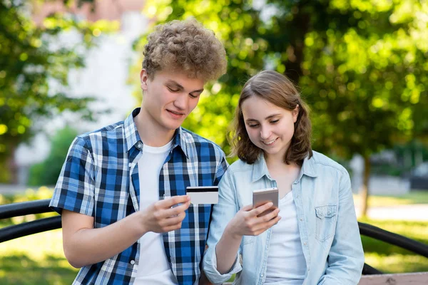 Una pareja joven, chico y chica. En verano en el parque en la naturaleza. Sostiene un smartphone en una tarjeta de crédito. Hacer compras en Internet. Sonríe feliz. Emocionalmente disfruta . —  Fotos de Stock