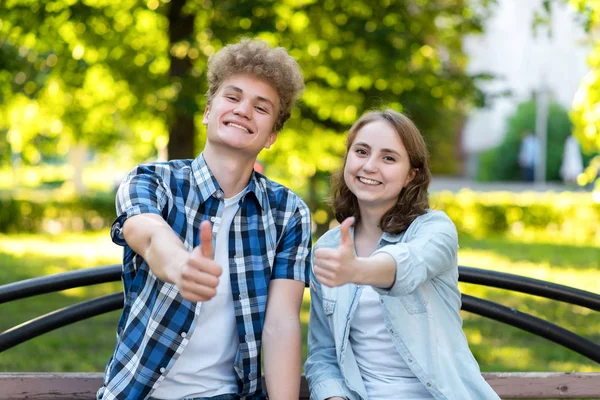 Молодая пара девочка и мальчик. Лето в парке на скамейке. Счастливо улыбается. Жесткие руки, типа, показывают большие пальцы вверх. Эмоциональные жесты. Концепция успеха и успеха . — стоковое фото
