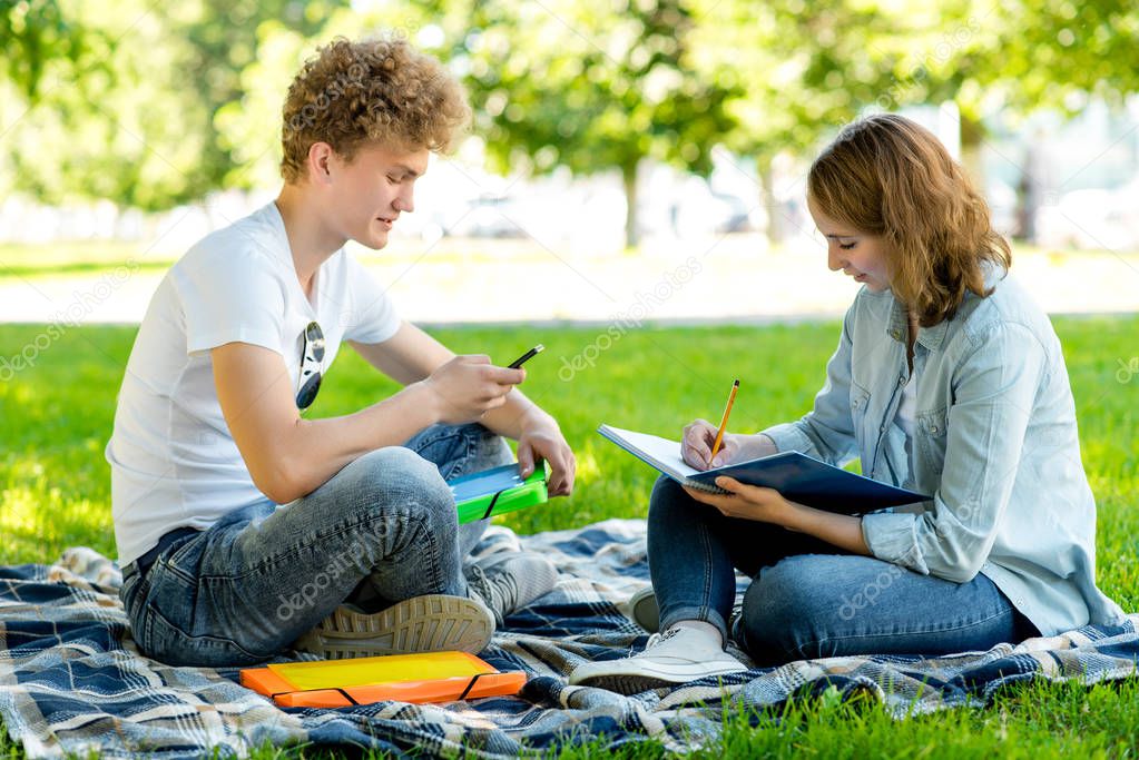 Students and schoolchildren in summer in the park. Girl and boy doing homework. In her hands holds a smartphone and a large notebook. Communicates makes notes in notepad.