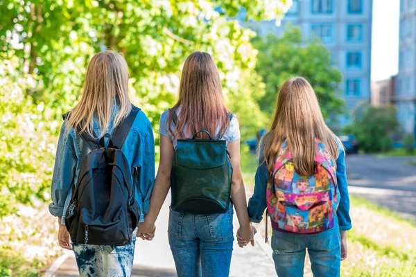 Три девочки-подростка. Они держат друг друга за руки. Возвращается домой после школы. Лето на природе. За рюкзаками. Вид сзади. Концепция школьных подружек . — стоковое фото