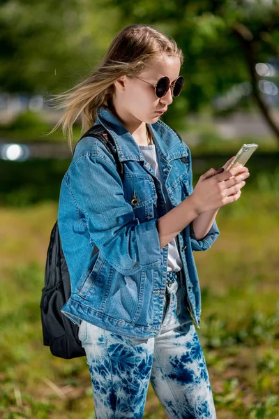 Seorang gadis remaja memegang smartphone. Musim panas di alam setelah sekolah. Mengenakan pakaian denim di belakang ranselnya. Menulis pesan kepada orang tua . — Stok Foto