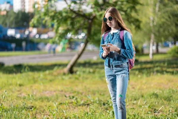 Gadis sekolahan. Musim panas di alam. Di tangan smartphone. Dia mengenakan pakaian jeans. Di belakang ransel. Ruang kosong untuk teks. Konsep komunikasi di Internet. Menulis pesan jaringan sosial . — Stok Foto