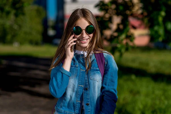 Seorang gadis remaja. Di musim panas sepulang sekolah. Di tangannya memegang smartphone. Dia berbicara di telepon. Selamat tersenyum. Dalam jeans sweater kacamata hitam . — Stok Foto