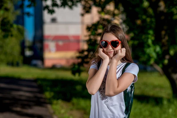 Une écolière. En été, après les cours. Derrière le sac à dos. Lunettes de soleil en forme de cœur. Espace libre pour le texte. Il pose positivement pour la caméra . — Photo