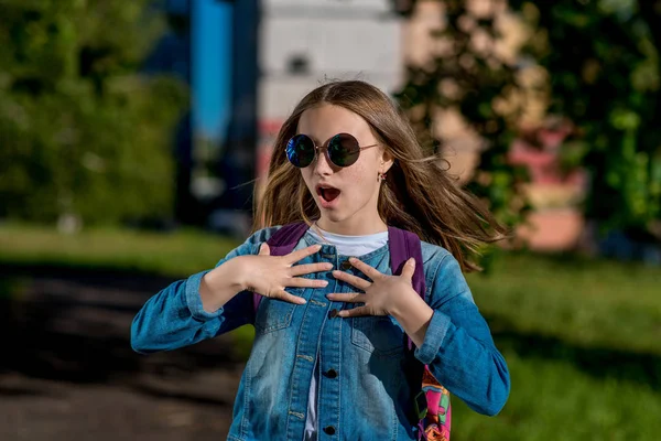 Девочка-школьница. Лето на природе. В джинсовой куртке с задней стороны рюкзака. Солнечные очки. Концепция неожиданного подарка. Эмоции счастья неожиданная радость . — стоковое фото