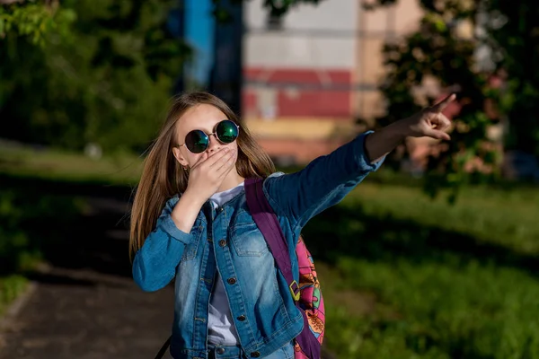 Девочка в джинсовой куртке, рюкзак в солнечных очках. Лето на природе. Она прикрывает рот ладонью. Палец указывает в сторону. Концепция неожиданности ужас случайности. Эмоции непонимания страха . — стоковое фото