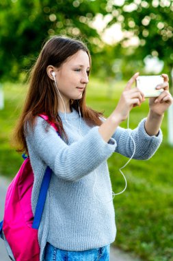 Güzel kız öğrenci kız Genç. Doğa yazın. Elinde bir smartphone çantasını arkasında tutar. Fotoğraflar telefon. Müzik kulaklık oynamak. Duygu mutlu bir şekilde gülümsüyor.