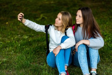 İki genç kız. Şehir Parkı yazında. Onlar çimlere oturup fotoğraf smartphone. Sırt çantaları. Okul dostluk kavramı. Duygu poz telefon.