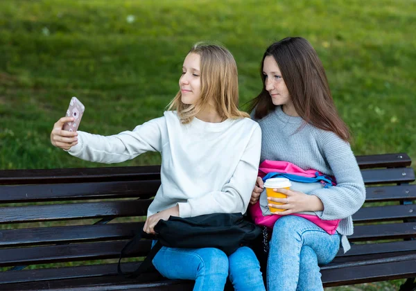 दोन लहान मुली शाळेत शिकत आहेत. उन्हाळ्यात खंडपीठावर. हाताने फोन ठेवला. स्मार्टफोनवर फोटो काढले. शाळेनंतर उन्हाळ्यात. सर्वोत्तम गर्लफ्रेंडची संकल्पना. भावना एक आनंद आहे . — स्टॉक फोटो, इमेज