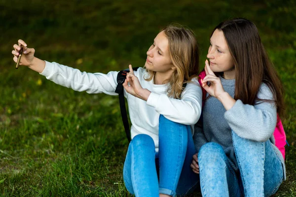 दोन अल्पवयीन मुली शहराच्या उन्हाळ्यात उद्यानात. ते गवत बसून स्मार्टफोनवर फोटो काढतात. बॅकपॅकच्या मागे. इंटरनेटवर व्हिडिओ कॉल करा. सामाजिक नेटवर्कमध्ये संप्रेषण . — स्टॉक फोटो, इमेज