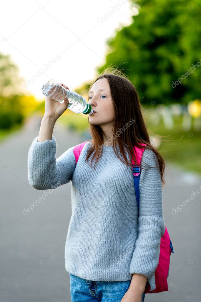 Beautiful school girl teenager. In summer in city park. He drinks water from the bottle. Behind the backpack. The concept of a healthy lifestyle. Emotions of happiness and enjoyment of pure water.