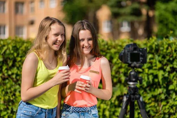 Dvě holky školačka. Léto v přírodě. Šťastně se usmívá. Ve svých rukou drží sklenici horké kávy nebo čaje. Píše video blog na kameru. — Stock fotografie