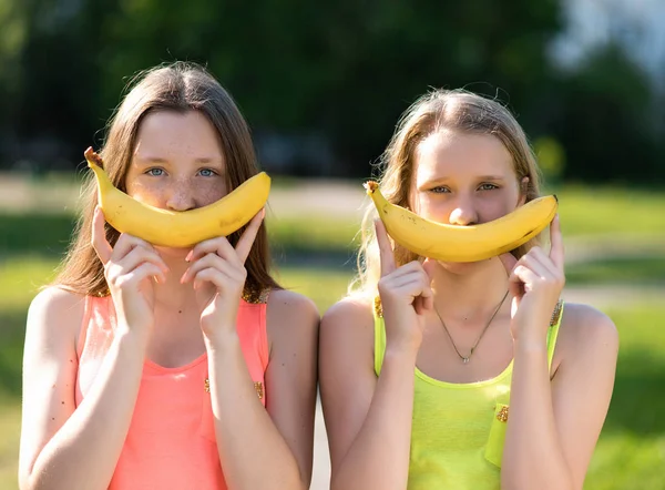 Dos colegialas. En sus manos tiene plátano. Verano en la naturaleza. El concepto de alimentación saludable. Las emociones son bromas y diversión . — Foto de Stock