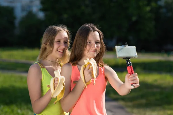 Dos chicas jóvenes. Jóvenes bloggers y bloggers. Sonríe feliz. Él sostiene el teléfono inteligente en sus manos, escribe vídeo en Internet. En las manos sosteniendo plátanos . — Foto de Stock