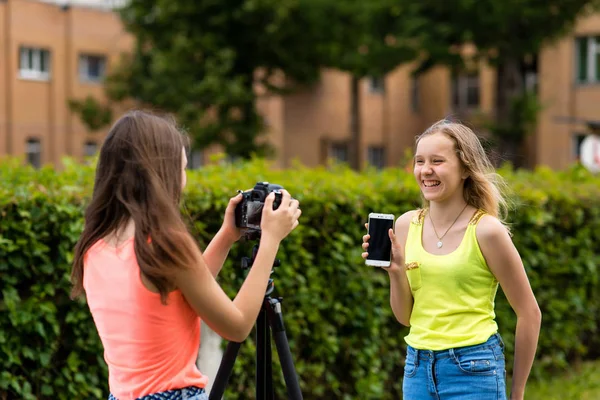 Τα δύο κορίτσια είναι οι έφηβοι. Το καλοκαίρι στη φύση. Γράφει το βίντεο με την κάμερα. Στα χέρια κρατά το τηλέφωνο. Έννοια αναθεώρηση smartphone. Χαμόγελα ευτυχώς. — Φωτογραφία Αρχείου