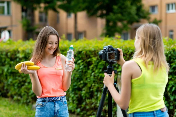 Dívky jsou školačky. Léto v přírodě. Emoční se šťastně usmívá. Zapíše na video kameru. V ruce drží láhev vody a banán. Koncept mladých bloggerů. — Stock fotografie