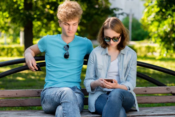 Un joven con una chica está sentado en un banco. En verano en el parque en la naturaleza. Manos sosteniendo un teléfono se comunica en las redes sociales. Vestido con ropa casual. Gafas de sol . —  Fotos de Stock