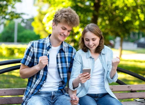 The guy with girl smiles happily. Emotion of winning large sums in the lottery. In his hands holds a smartphone and a credit card. In summer, on a park bench. The concept of success and good luck.