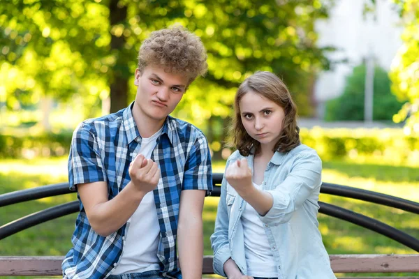 Un tipo con una chica en el verano en el parque en un banco. El gesto de las manos muestra una figura. El concepto de desconfianza. Emociones terribles. Estudiantes, renuencia a dar. Manos mostrando puños . —  Fotos de Stock