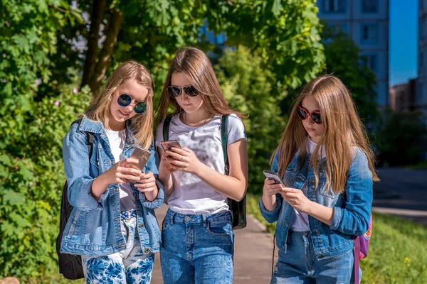 Три девочки школьницы. Лето на улице. В его руках смартфон. В джинсовой одежде, солнечных очках. Концепция детей в социальных сетях. Написать сообщение в Интернете . — стоковое фото