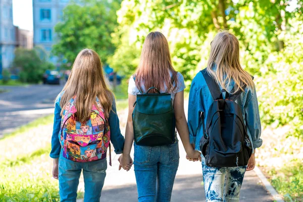 Tři kamarádky holka školačka. Drží se navzájem ruce. Léto ve městě. Pohled zezadu. Do školy chodí po ulici. Batohy za zády. Koncept je nejlepší přátelé. — Stock fotografie