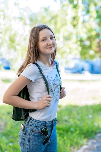 Colegiala detrás de la mochila. Verano en la naturaleza. Posando en cámara. Ella sonríe emocionalmente . — Foto de Stock