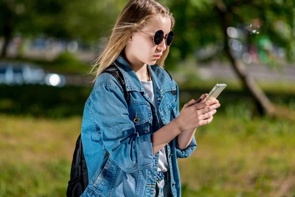 夏用デニムの服で 10 代の少女。後ろのサングラスのバックパック。彼の手でスマート フォンを保持します。インターネット上の通信の概念。スマート フォン メッセージを書き込みます. — ストック写真
