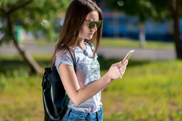 Een tienermeisje in de zomer in denim kleding. Achter de rugzak in zonnebril. In zijn handen houdt een smartphone. Concept leest berichten op sociale netwerken op een smartphone. — Stockfoto