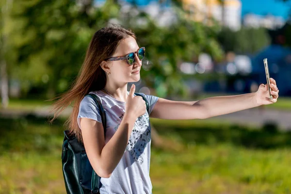 Krásná brunetka. V létě školačka má přestávku. Ve svých rukou drží smartphone. Zahájení audiovizuálního volání. Vyfoťte sami sebe na telefonu. — Stock fotografie