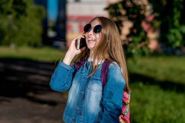 Gadis sekolahan berbicara tentang smartphone. Pada musim panas, dalam pakaian denim, ransel dengan kacamata hitam berada di belakangnya. Dia tertawa terang dan emosional. Senyum dan nikmati percakapan . — Stok Foto