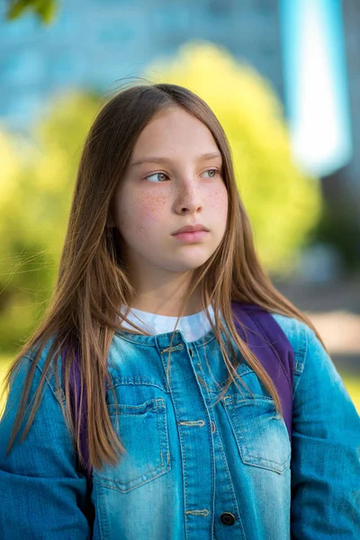 Close-up portrait of a schoolgirl girl. Summer in the city on nature. Somewhere in jeans clothes behind backpack. Long hair on face freckles. Emotional look aside. — Stock Photo, Image