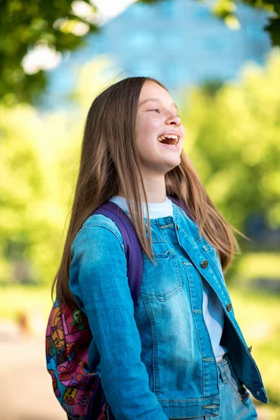 Hermosa colegiala. Verano en la naturaleza. Vestida con ropa vaquera detrás de su mochila. Concepto de vuelta a la escuela. Emoción de felicidad placer placer, niños brillantes risa . — Foto de Stock