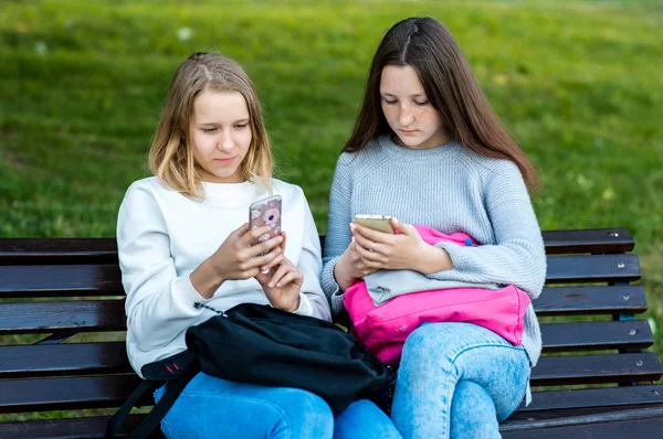 二人の女の子が女子学生です。夏に、彼らは自然の中のベンチに座っています。スマート フォンを押しの手で書き換えのメッセージを読み取ります。インターネット上で子供のコンセプトです。放課後休憩します。. — ストック写真