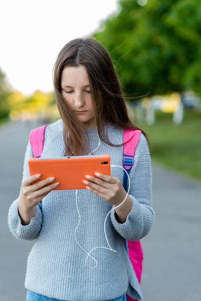 女の子女子高生のティーンエイ ジャー。自然の中の夏。彼女の手の中には、タブレットを保持しています。音楽を聴いたり、ヘッドフォンで映画を見る。放課後の休暇上の子供の概念。バックパックの後ろに. — ストック写真