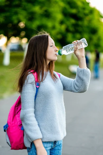 Девочка-школьница. Лето в городе на улице. Он пьёт воду из бутылки. За рюкзаком. Концепция обеда после школы. Эмоции жаждут удовольствия . — стоковое фото