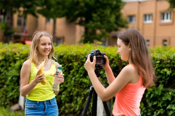 Twee meisjes schoolmeisje. In de zomer in de stad. Schrijft de video naar de camera. In de handen houdt bananen en een fles water. Het concept van een video blog over gezond eten. — Stockfoto