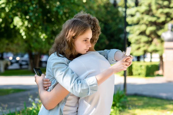 The girl hugs a guy in summer in nature. In the hands of holding smartphones communicates in social networks. Students after school rest. Happy beginning of the relationship. Best friends forever. — Stock Photo, Image