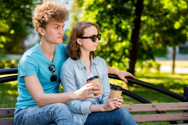 Giovane bella coppia di studenti. Estate in parco nella natura. Si siede sulla panchina. Tiene in mano delle tazze di caffè. Con gli occhiali da sole. Gode di relax dopo la scuola . — Foto Stock