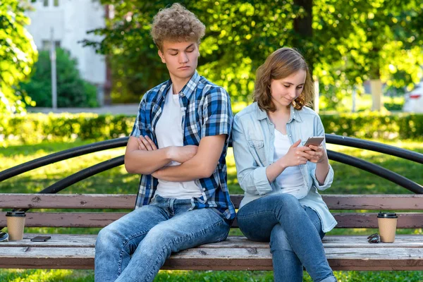 The guy and the girl are sitting on bench. The girl writes messages in social networks. The guy spies on correspondence. The concept of distrust in a relationship. Family problems.