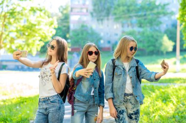 Üç genç kız. Doğa yazın. Elinde smartphone tutar. Telefonda fotoğraf çekmek. Güneş gözlüğü ve denim giysiler. Kendi telefon kavramı. Duygu mutlu bir şekilde gülümsüyor.