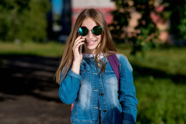 Gadis kecil sekolahan remaja. Di musim panas di taman alam. Bicara di telepon. Dengan pakaian jins dan kacamata hitam. Emosi kebahagiaan sukacita dan tersenyum . — Stok Foto