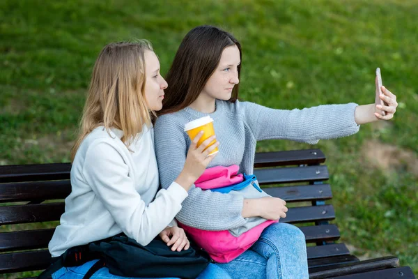 Malé školačky sedí na lavičce. V létě v parku jsou 2 holky. Kemp. V rukou drží sklenku kávy. Fotit sebe na telefonu. — Stock fotografie