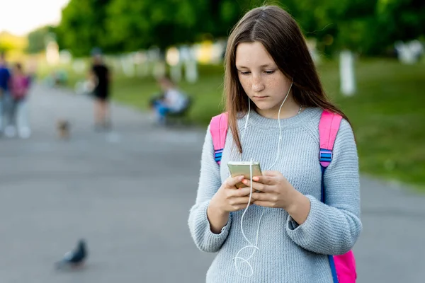 Meisje schoolmeisje tiener in de zomer in het park buiten. In haar handen houdt een smartphone luisteren naar muziek op koptelefoon, het schrijven van berichten op de telefoon. Vrije ruimte voor tekst. — Stockfoto