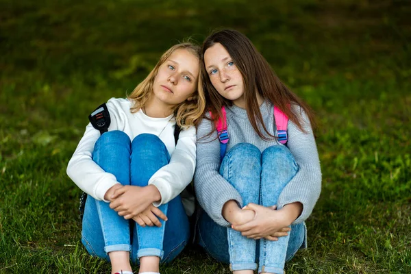 Two girls schoolgirl sitting in the summer on grass in the park. Emotions of sadness and sadness. The concept is best friends, girlfriends.