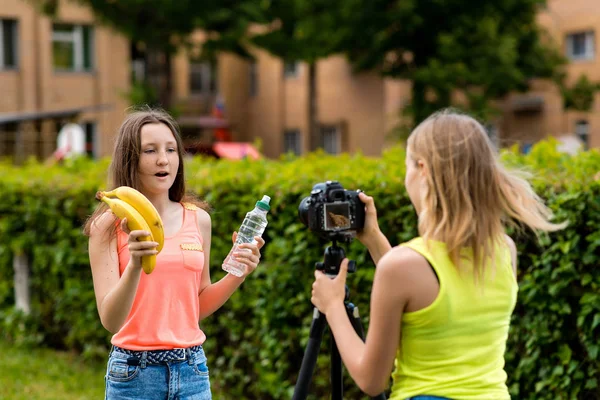 Νεαρά κορίτσια bloggers, το καλοκαίρι στην πόλη στη φύση. Εγγραφή βίντεο στη φωτογραφική μηχανή. Στα χέρια της κρατά ένα μπουκάλι νερό και δύο μπανάνες. Η έννοια της υγιεινής διατροφής. — Φωτογραφία Αρχείου