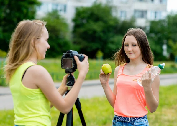 2 kız gençler vardır. Doğa parkında yazın. Video kamera için yazar. Heres nasıl bir elma ve bir şişe su tutar. Genç blogcular kavramı. Kullanma fotoğraf makinesi tripod ile. — Stok fotoğraf