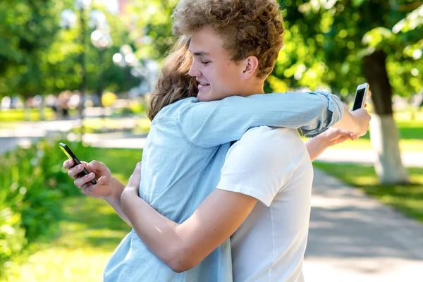 Chlap je objímat dívku v létě v parku. Šťastný úsměv držel ruku s chytrými telefony. Citově navzájem důvěřují. Silné objetí navzájem. Studenti po institutu na dovolené. — Stock fotografie