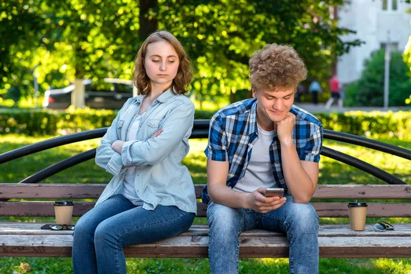 Un chico y una chica están sentados en un banco. El tipo está escribiendo un mensaje en las redes sociales. Una chica está espiando por correspondencia. Conceptos de desconfianza en las relaciones. El problema en la familia . —  Fotos de Stock