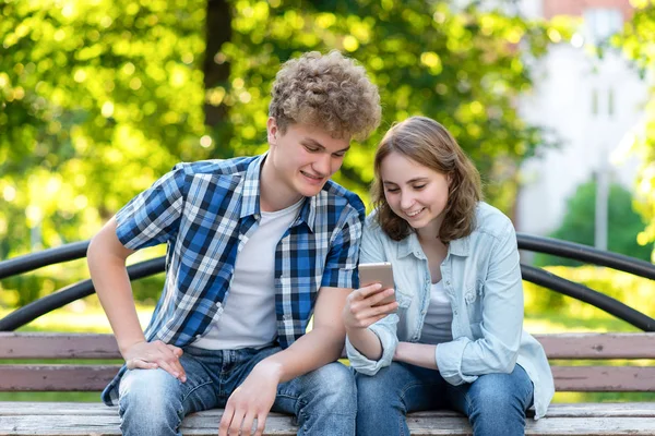 Mladý pár v létě v parku venku. Sedí na lavičce v městě. Šťastně se usmívá. V rukou drží smartphone umožňuje nákupy na internetu. Kemp. Koncept šťastné rodiny. — Stock fotografie