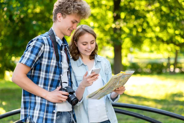Mladý kluk s holkou. V létě v městě v přírodě. Turisté se drží cestovní mapu. V rukou chytrý telefon a fotoaparát. Cestuje do aplikace na telefonu. — Stock fotografie
